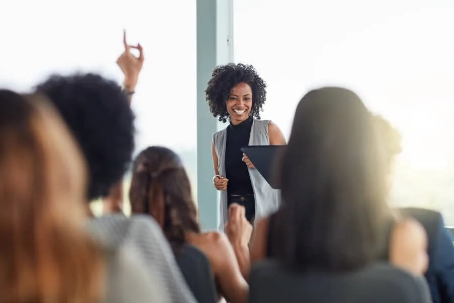 Smiling woman leading a session or workshop, with audience members raising their hands