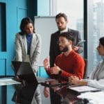 A sales metrics meeting where four people are discussing beside a laptop