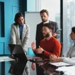 A sales metrics meeting where four people are discussing beside a laptop